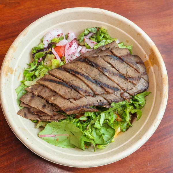 Grilled meat slices over a fresh green salad with red onions and tomatoes in a beige bowl.