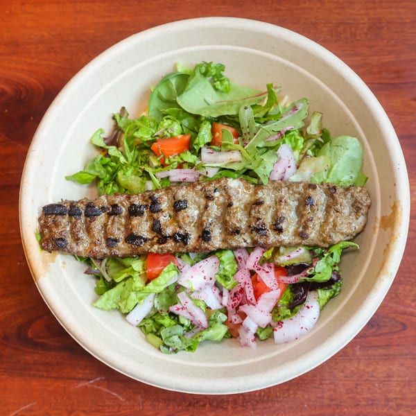 Grilled kofta kebab on a bed of fresh mixed greens, tomatoes, and red onions in a bowl.