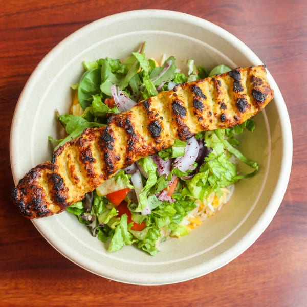 Grilled chicken skewer over a bed of rice and fresh salad in a round bowl.